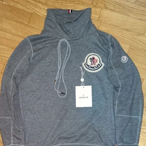 Grå polotröja från Moncler - Säljer en grå polotröja från Moncler med stor logotyp på bröstet och mindre logga på ärmen. Tröjan har långa ärmar och hög krage med dragsko. Perfekt för kyligare dagar och riktigt snygg till jeans.