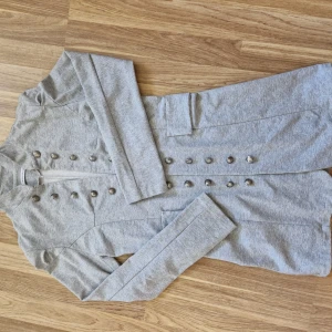 Grå kavaj med dekorativa knappar - Säljer en snygg grå kavaj med hög krage och dubbla rader dekorativa knappar framtill. Kavajen har lång ärm, markerade axlar och två fejk fickor framtill. Perfekt för dig som vill ha en stilren och trendig look.