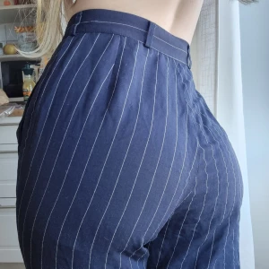 Mörkblå randiga kostymbyxor - Säljer ett par mörkblå kostymbyxor med vita tunna ränder. Byxorna är högmidjade med normal passform och har knäppning fram. Perfekta för en stilren och trendig look.