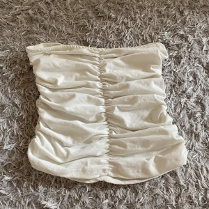 Vit tubtopp med rynk från NA-KD - Säljer en vit tubtopp från NA-KD med snygga rynkdetaljer framtill. Toppen är axelbandslös och har en glansig finish som ger en stilren look. Perfekt att matcha med jeans eller kjol.