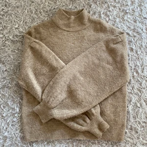 Beige stickad polotröja från Vero Moda - Mysig beige stickad polotröja från Vero Moda med ribbad struktur. Tröjan har långa ärmar med breda muddar och en hög krage som ger en stilren look. Perfekt för lager-på-lager under kyliga dagar. Lappen är bortklippt men tröjan ska tvättas i 30 grader med liknande färger. 