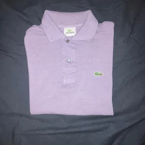 Lila pikétröja från Lacoste knappt använd - Säljer en klassisk lila pikétröja från Lacoste i 100% bomull. Tröjan har krage, två knappar framtill och den ikoniska gröna krokodilloggan på bröstet. Perfekt för en stilren och avslappnad look. 