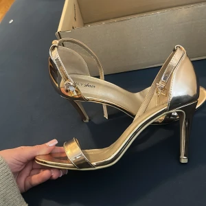 Guldiga klackskor från NLY Shoes - Säljer ett par glansiga guldiga klackskor från NLY Shoes. Skorna har öppen tå, smal rem över tårna och runt vristen samt hög klack. Perfekta för att lyfta vilken outfit som helst.