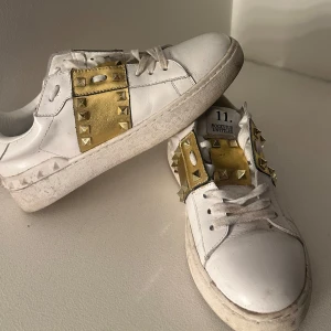 Vita sneakers med guldiga nitar  - Säljer ett par vita (icke äkta) Valentino sneakers med bred guldfärgad rem och stora guldiga nitar över vristen. Skorna har klassisk rund tå och platt sula. Perfekta för dig som vill sticka ut med en lyxig och edgy detalj.