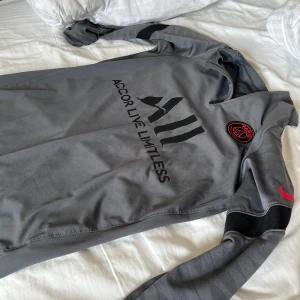Grå långärmad Paris Saint-Germain fotbollströja - Säljer en grå långärmad PSG-tröja från Nike med svart tryck 'ALL Accor Live Limitless' på bröstet och 'Visit Rwanda' på ryggen. PSG-emblem i rött och svart på bröstet samt röd Nike-logga på ärmen. Tröjan har dragkedja vid halsen och diskret mönster på ärmarna.