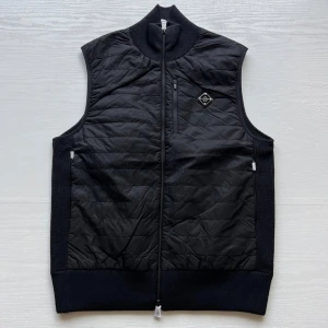 Svart quiltad väst från J.Lindeberg - Stilren svart väst från J.Lindeberg med quiltat mönster och hel dragkedja framtill. Västen har ribbade partier vid krage, sidor och nederkant samt en bröstficka med dragkedja och diskret logomärke på bröstet. Pris kan diskuteras. Skick 10/10, helt oanvänd, storlek M och kostar egentligen 2499 kr. Kontakta vid mer information.