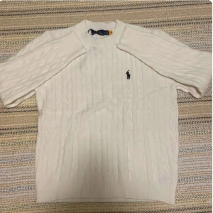 Ralph lauren ull tröja  - Vit  och grå kabelstickad tröja från Ralph Lauren med rund halsringning och klassisk broderad logga på bröstet. Tröjan har lång ärm och ribbade muddar vid ärmslut och nederkant. Perfekt för en stilren look.