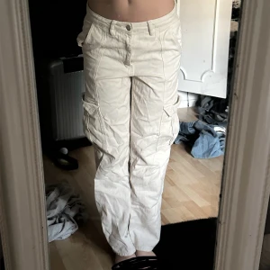 Vita cargopants med fickor - Säljer ett par vita cargopants med flera stora fickor på sidorna och raka ben. Byxorna har normal passform och stängs med knapp och dragkedja. Perfekta för en avslappnad och trendig stil.