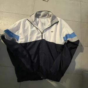 Vit och marinblå vindjacka från Lacoste - Säljer en snygg vindjacka från Lacoste i vitt och marinblått med blå detalj på ärmarna. Jackan har dragkedja framtill, hög krage och klassisk Lacoste-logga på bröstet. Perfekt för en sportig och stilren look. Väldigt bra skick, pris kan diskuteras vid snabb affär!