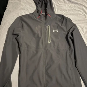 Under Armour jacka - Säljer en stilren svart vindjacka från Under Armour med huva och dragkedja. Jackan har en praktisk bröstficka med dragkedja och är perfekt för utomhusaktiviteter. Den har långa ärmar och en sportig look.