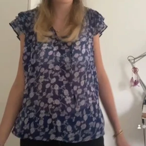 Blommig blus i blått och vitt - Säljer en söt blommig blus i blått och vitt. Blusen har korta ärmar och ett luftigt fall, perfekt för varmare dagar. Den har en liten v-ringning och är gjord i ett lätt material.