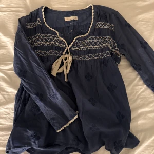 Blå blus med broderade detaljer - Charmig blå blus med vita broderade detaljer och knytband framtill. Blusen har långa ärmar och ett diskret blommönster. 