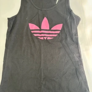 Svart  linnetopp från Adidas - Säljer en svart linnetopp från Adidas med rosa logga framtill. Perfekt för träning eller en avslappnad dag. Linnet är ärmlöst och har en rund halsringning.