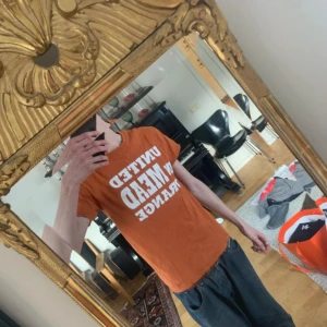 Orange t-shirt med tryck - Säljer en orange t-shirt med texten 'UNITED IN MEAD ORANGE' på framsidan och 'WE ARE MEAD' på baksidan. T-shirten har en klassisk passform och korta ärmar.