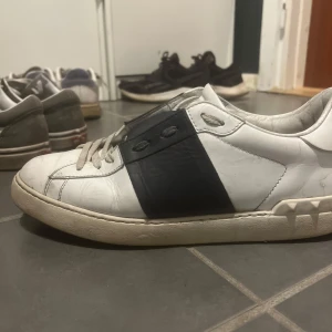 Valentino Garavani sneakers med svart detalj - Snygga Valentino Garavani sneakers i vitt skinn med en bred svart rem över snörningen. Skorna har en unik design med nitar runt hälen och klassisk snörning framtill. Perfekta för en stilren look.