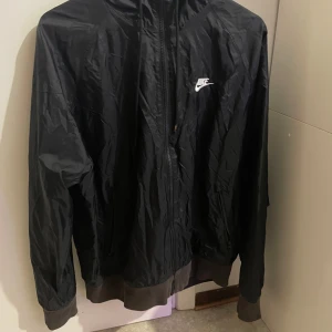 Svart vindjacka från Nike - Säljer en svart vindjacka från Nike med dragkedja och huva. Jackan har långa ärmar och en vit Nike-logga på bröstet. Perfekt för blåsiga dagar.