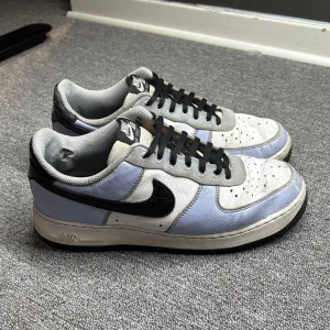 Nike Air Force 1 custom made on Nike.com - Säljer ett par klassiska Nike Air Force 1 sneakers i vitt och ljusblått med svarta detaljer. Skorna har en ikonisk design med perforerad tå och svart swoosh. Perfekta för en stilren look.
