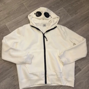 Vit hoodie med glasögon i huvan - Säljer en unik vit hoodie från C.P. Company med inbyggda glasögon i huvan. Den har en dragkedja framtill och långa ärmar. Perfekt för en cool och avslappnad stil.