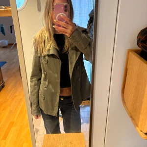 Grön trenchcoat - Säljer en snygg grön trenchcoat med dubbelknäppning, passar perfekt för våren!