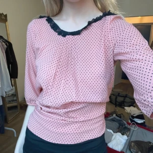 Rosa prickig blus med spetsdetalj - Säljer en söt rosa blus med små svarta prickar och spetsdetalj vid halsringningen. Blusen har trekvartsärmar och en bekväm passform. Perfekt för en stilren look!