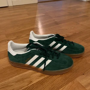 Gröna Adidas Gazelle i mocka - Säljer ett par snygga gröna Adidas Gazelle i mocka med vita detaljer och snörning. Skorna har en klassisk design med tre vita ränder på sidan och en gummisula i brunt. Perfekta för en stilren look. Använda endast en gång för en photoshoot.