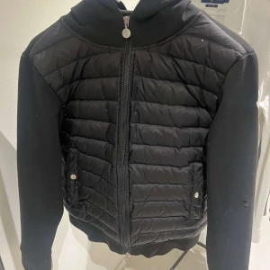 Svart quiltad jacka från Moncler - Snygg svart quiltad jacka från Moncler med dragkedja och långa ärmar. Jackan har en stilren design med ribbade muddar och två fickor med knappar framtill. Perfekt för kyligare dagar men även våren.