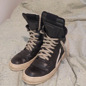 2011 Rick Owens Geobaskets - Säljer ett par coola höga sneakers från Rick Owens i svart och vitt. Skorna har snörning och dragkedja på sidan för en unik look. Perfekta för dig som vill ha något stilrent men ändå edgy.