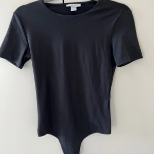 Svart kortärmad bodysuit - Snygg och enkel svart bodysuit med korta ärmar. Perfekt för att matcha med jeans eller kjol för en stilren look. Den har en rund halsringning och är gjord i ett mjukt material för bekväm passform.
