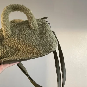 Grön väska i teddymaterial med axelrem - Säljer en mysig grön väska i teddymaterial med både handtag och justerbar axelrem. Väskan har en dragkedja upptill och flera fack inuti för enkel organisering. Perfekt för att ge din outfit en unik touch!