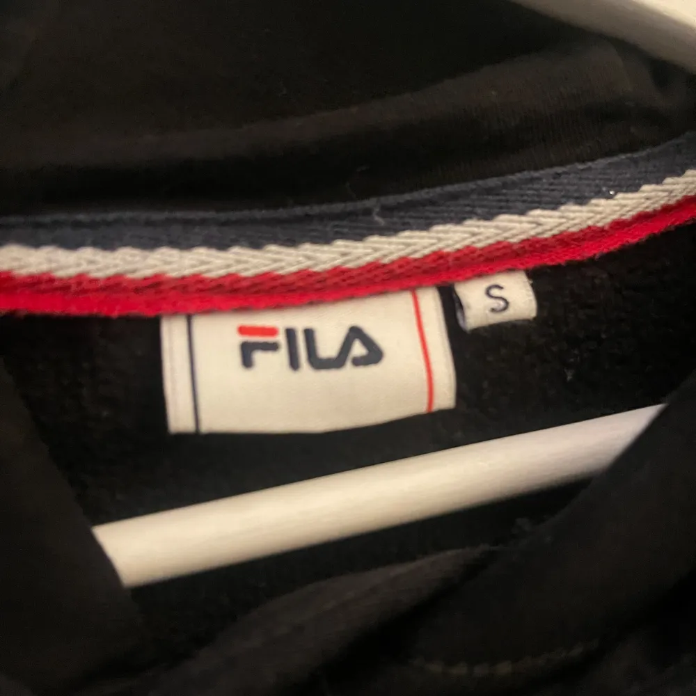 Säljer en svart hoodie från Fila med deras logga i rött och vitt på framsidan. Tröjan har en klassisk design med huva och dragsnören samt en stor ficka framtill. Perfekt för en avslappnad stil.. Neuletakit & Villapaidat.