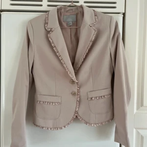 Beige/rosa kavaj från H&M - Säljer en elegant beige/rosa kavaj från H&M med volangdetaljer längs kragen, fickorna och nederkanten. Kavajen har två knappar framtill och långa ärmar. Perfekt för en stilren look. Den är lite för liten för mig! Men älskar modellen och färgen! 