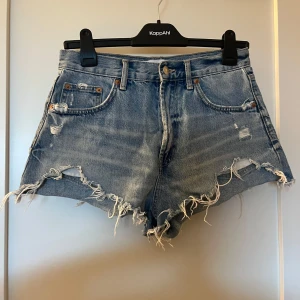 Blå jeansshorts från Zara - Säljer ett par snygga blå jeansshorts från Zara med fransiga kanter och slitna detaljer. Perfekta för en avslappnad sommarlook. De har en klassisk femficksdesign och knappgylf. Passar perfekt för varma dagar!