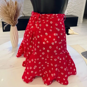 Röd blommig omlottkjol - Charmig röd omlottkjol med vita blommor och volangdetaljer. Kjolen har en knytning i midjan som ger en fin passform och en lekfull look. Perfekt för sommarens alla tillfällen. Passar perfekt i s/m/l storlek. Jag rekommenderar den varmt.🌸👌