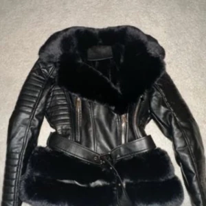 Svart skinnjacka med pälsdetaljer - Snygg svart skinnjacka med mjuka pälsdetaljer och bälte i midjan. Jackan har dragkedjor och quiltade detaljer på ärmarna. Perfekt för en stilren look.