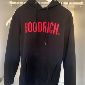 Svart hoodie från Hoodrich - Säljer en svart hoodie från Hoodrich med röd text på framsidan. Den har en klassisk design med huva och dragsko samt en stor ficka framtill. Perfekt för en avslappnad stil.