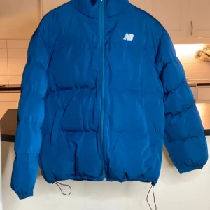 Blå pufferjacka från New Balance - Säljer en blå pufferjacka från New Balance med dragkedja och hög krage. Jackan har en normal passform och är perfekt för kyligare dagar. Den har långa ärmar och justerbar nederkant för extra komfort.