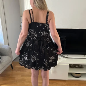 Svart klänning med stjärnmönster från ONLY - Snygg svart klänning från ONLY med ett coolt stjärnmönster. Klänningen har en v-ringad design och smala axelband. Perfekt för en kväll ute eller en speciell tillställning. Den har en knytning i midjan för en smickrande passform.