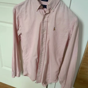 Rosa skjorta från Polo Ralph Lauren - Säljer en stilren rosa skjorta från Polo Ralph Lauren. Skjortan är tillverkad i 100% bomull och har långa ärmar samt knappar framtill. Den klassiska loggan är broderad på bröstet. Perfekt för en snygg och avslappnad stil.