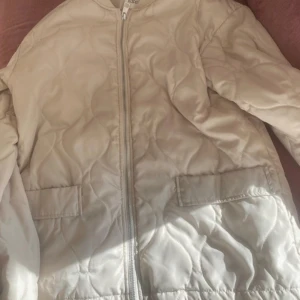 Beige /crèmefärgad quiltad jacka från Kappahl - Säljer en  beige quiltad jacka från Kappahl. Jackan har en dragkedja framtill och finna fickor. Perfekt för kyligare dagar med sin långa ärm och quiltade design.