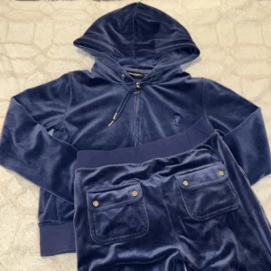Mörkblå velourset från Juicy Couture - Säljer ett snyggt mörkblått velourset från Juicy Couture. Setet består av en hoodie med dragkedja och byxor med snörning i midjan. Perfekt för en avslappnad stil med en touch av lyx. 💙