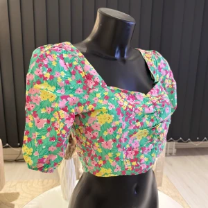 Blommig topp med puffärmar - Säljer en färgglad blommig topp med puffärmar och rynkdetaljer framtill. Toppen har en fyrkantig urringning och är perfekt för sommarens alla tillfällen. Den har en härlig mix av grönt, rosa, gult och rött. Passar parfekt i S/M/L storlek.
