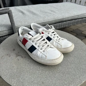 Vita sneakers från Moncler - Snygga vita sneakers från Moncler med klassiska ränder i rött och blått på sidan. Skorna har snörning och en rund tå, vilket ger en stilren look. Perfekta för en casual stil med en touch av lyx. Kolla sista bilden för en liten skada på ena skon. Hör gärna av er vid frågor eller annat! (köpta på vestiare)
