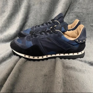 Valentino Garavani sneakers i blått och svart - Snygga sneakers från Valentino Garavani i en cool blå och svart färgkombination med camouflage-mönster. Skorna har dekorativa nitar runt sulan och är tillverkade i en mix av mocka och syntetmaterial. Perfekta för att ge din stil en edgy touch.