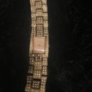 Guldfärgad klocka från D&G - Elegant guldfärgad klocka från D&G med glittrande stenar på armbandet och urtavlan. Perfekt för att lägga till en touch av glamour till din stil. Klockan har en rektangulär urtavla och ett detaljerat armband.