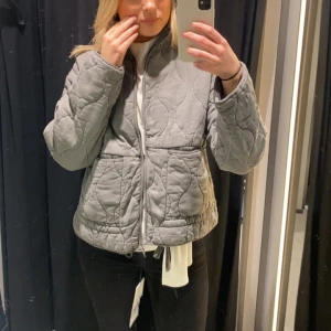 Grå quiltad jacka - Säljer en stilren grå quiltad jacka med dragkedja och hög krage. Jackan har en unik vågig quiltning och två stora fickor framtill. Perfekt för våren💗