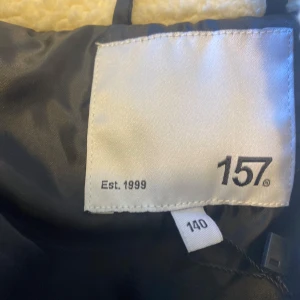 Vit teddyjacka från 157 - Mysig vit teddyjacka från 157 helt ny utan prislapp med svart dragkedja och en praktisk bröstficka. Perfekt för kyliga dagar med sin mjuka och varma känsla. Jackan har långa ärmar och en stilren design.