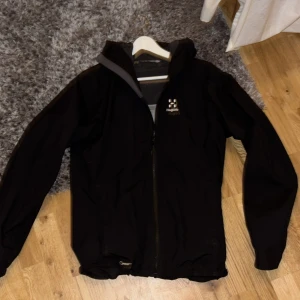 Svart vindjacka från Haglöfs - Säljer en svart vindjacka från Haglöfs med dragkedja och huva. Jackan har ett litet logotypmärke på bröstet och är perfekt för blåsiga dagar. Den är funktionell och stilren, idealisk för utomhusaktiviteter. Även regnskyddad