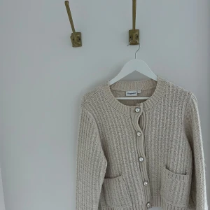 Beige stickad kofta  - Elegant beige stickad kofta med fina pärlknappar och två praktiska fickor framtill. Perfekt för en stilren look. Storlek L men är mer som en M.