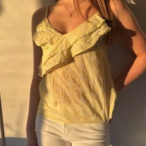 Gult linne - Säljer en söt gul linnetopp med broderade blommor och volangdetaljer. Toppen har smala axelband och en v-ringad design. Perfekt för soliga dagar!