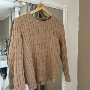 Beige stickad tröja från Polo Ralph Lauren - Säljer en snygg beige stickad tröja från Polo Ralph Lauren med kabelstickat mönster. Tröjan har en klassisk rund halsringning och långa ärmar. Perfekt för kyligare dagar!